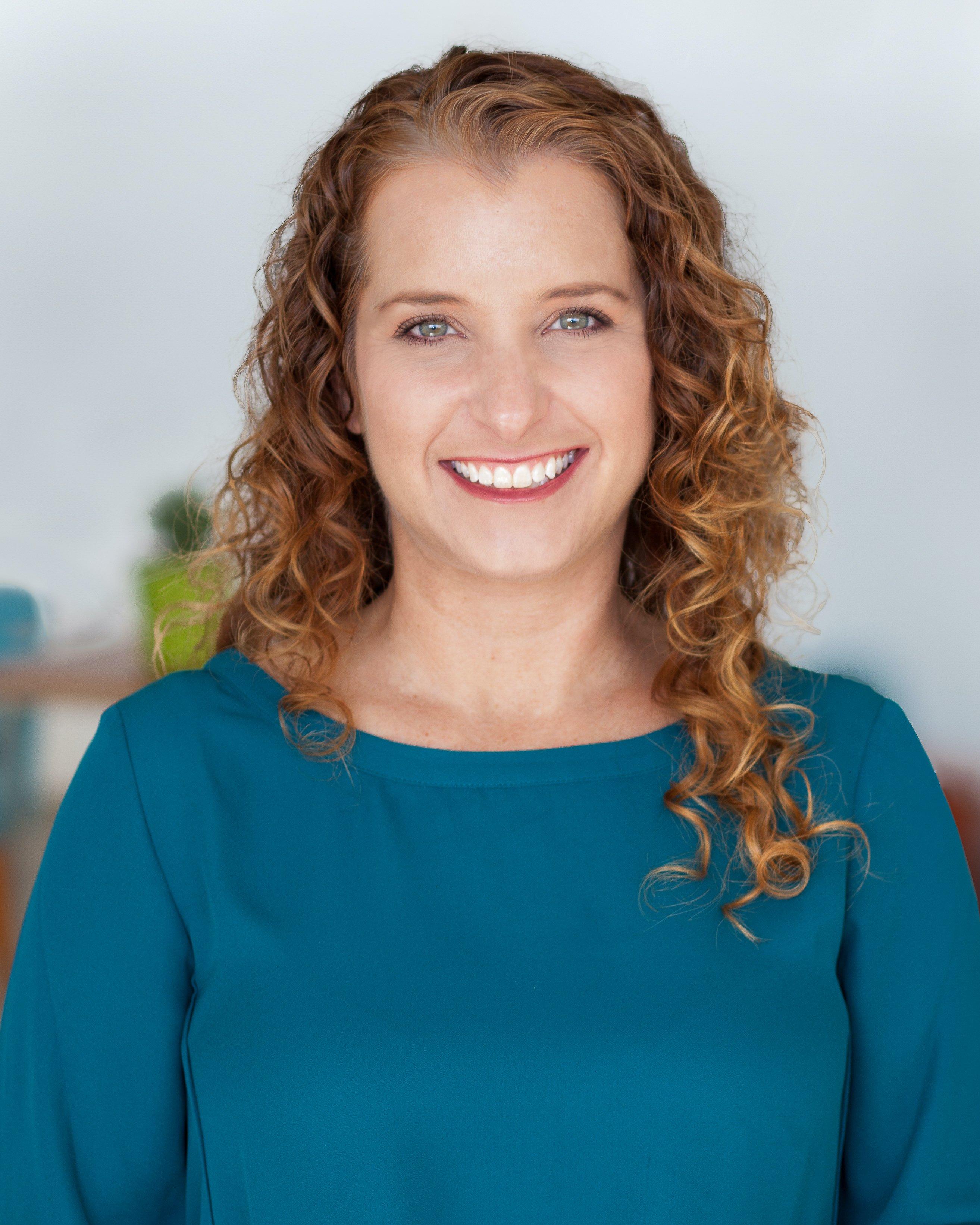 Professional headshot of a person with curly brown hair wearing a teal dress, smiling warmly against a light background