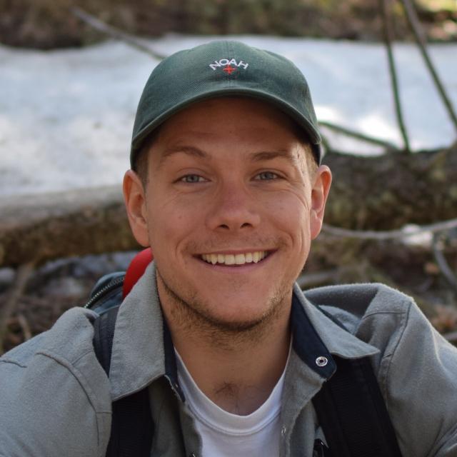 Close-up portrait of a smiling person wearing a green Noah baseball cap and gray jacket outdoors