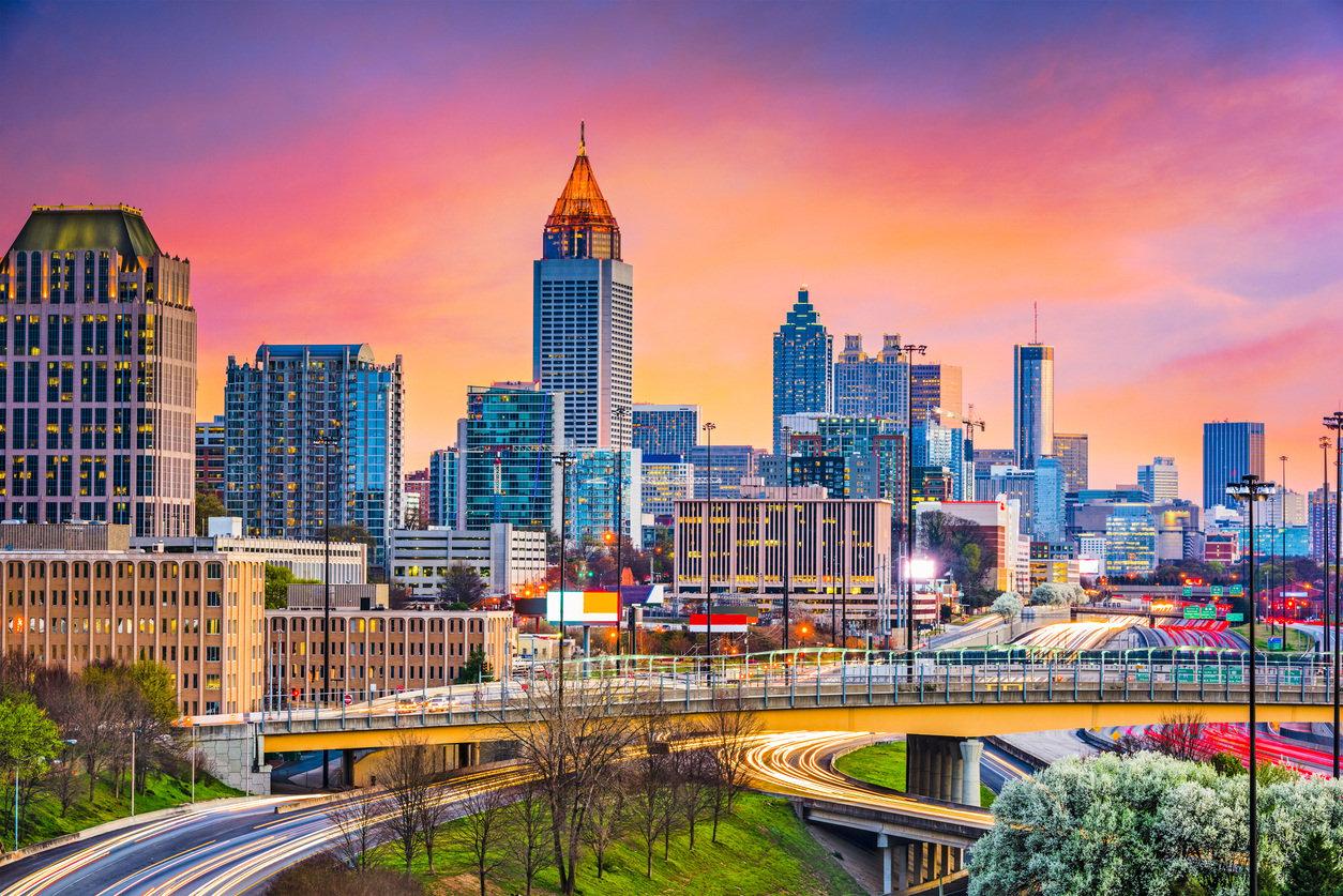 Downtown Atlanta skyline with illuminated skyscrapers against a vibrant pink and orange sunset sky, with highway light trails below