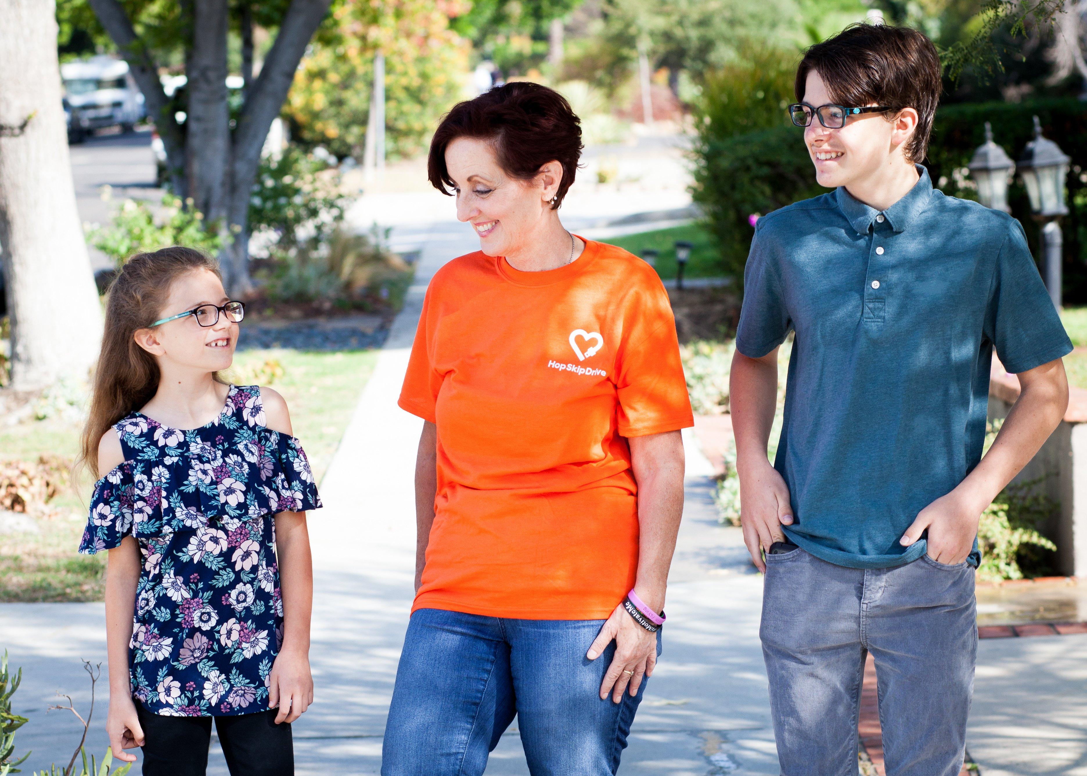 Three people smiling outdoors, with woman in orange HopSkipDrive shirt standing between two younger individuals in casual attire