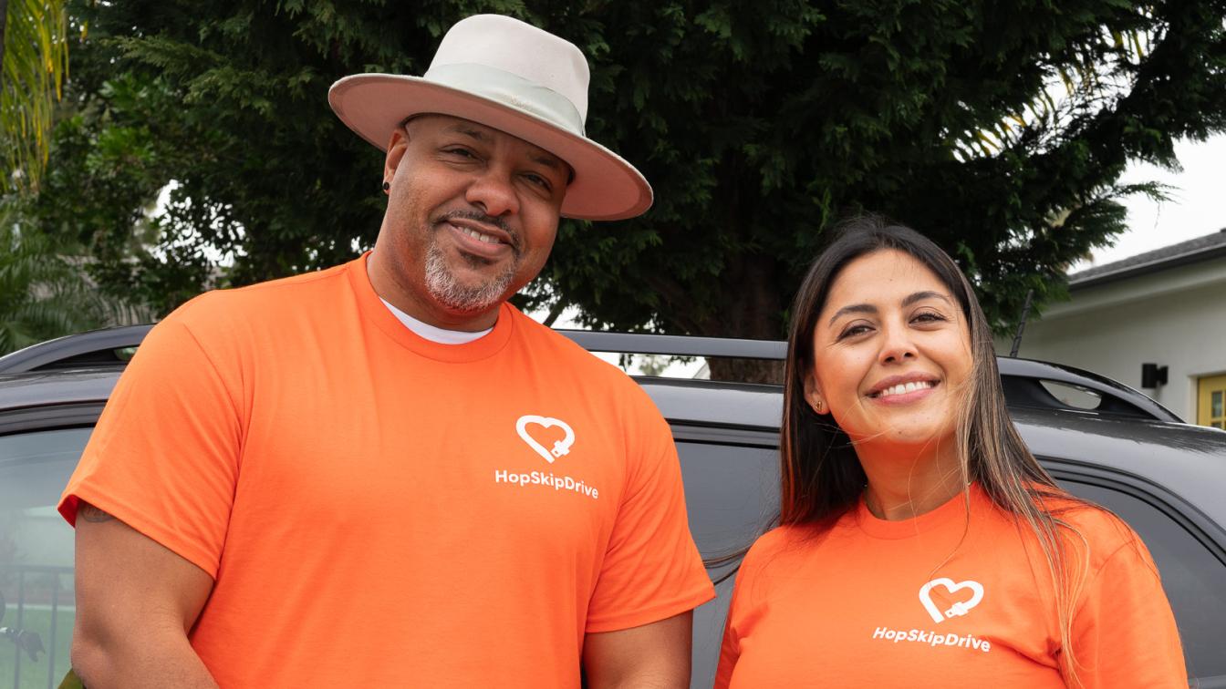 Two team members wearing bright orange HopSkipDrive t-shirts with heart logo standing in front of a car, one wearing a white hat