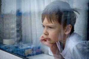 Child resting head on hand while thoughtfully gazing through a rain-spotted window, with blurred city background