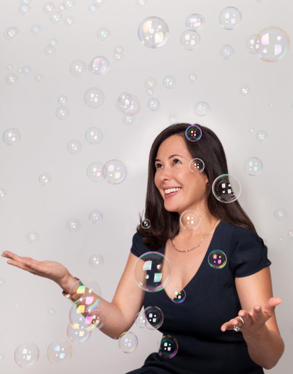 Woman in black dress smiling while surrounded by floating iridescent soap bubbles against light background