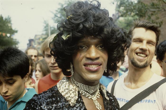 A close-up portrait of a person in a black sequined outfit with curly black wig and rhinestone jewelry, surrounded by festival attendees