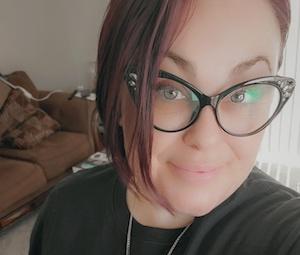 Close-up portrait of a person with cat-eye glasses, short dark hair, and a gentle smile wearing a dark top