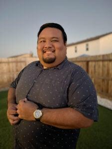 Person smiling warmly at camera in backyard setting during golden hour, wearing blue polka dot shirt