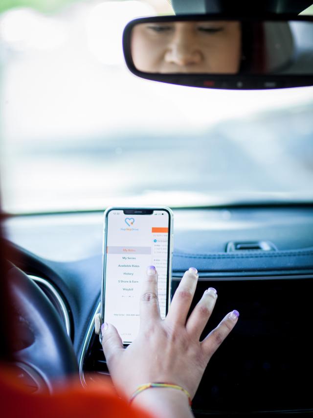 Hand reaching for smartphone mounted on car dashboard displaying a mobile app interface, with rearview mirror visible above