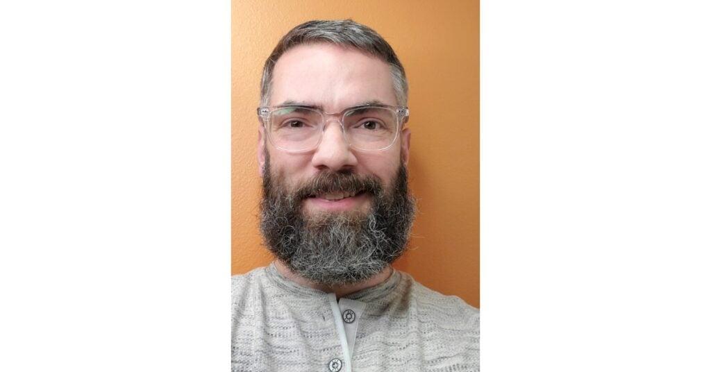 Professional headshot of person wearing clear-framed glasses and full beard, smiling warmly against an orange wall