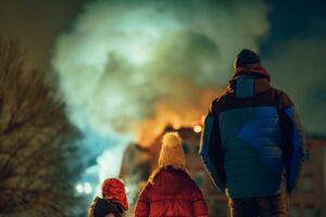 Three people in winter coats watching a bonfire with smoke illuminated against dark night sky