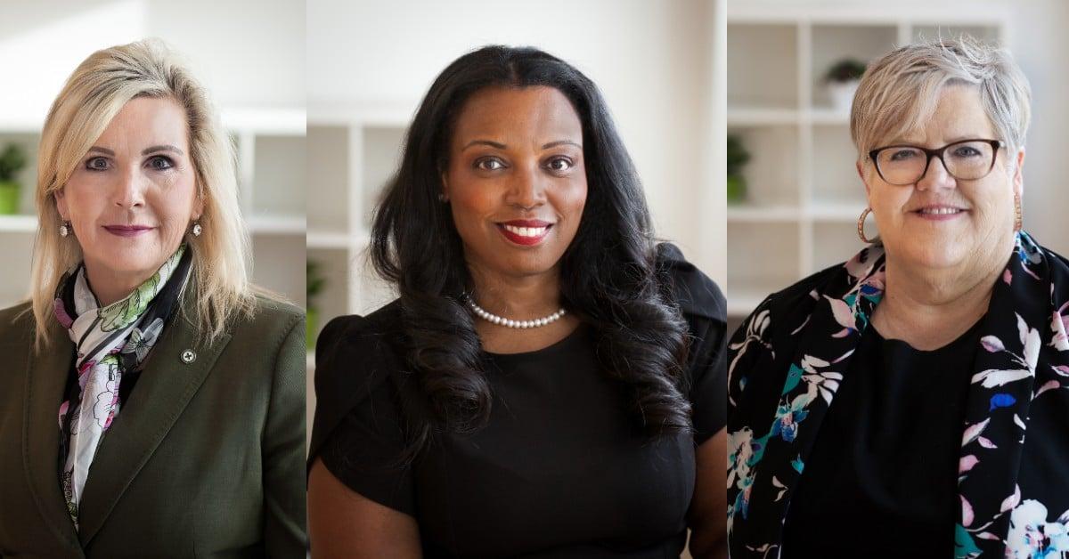 Three professional women in business attire posed for corporate headshots against a light background with built-in shelving