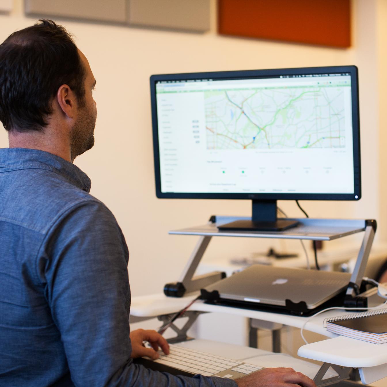 A professional in a blue shirt working at a standing desk setup, viewing digital maps on a large monitor