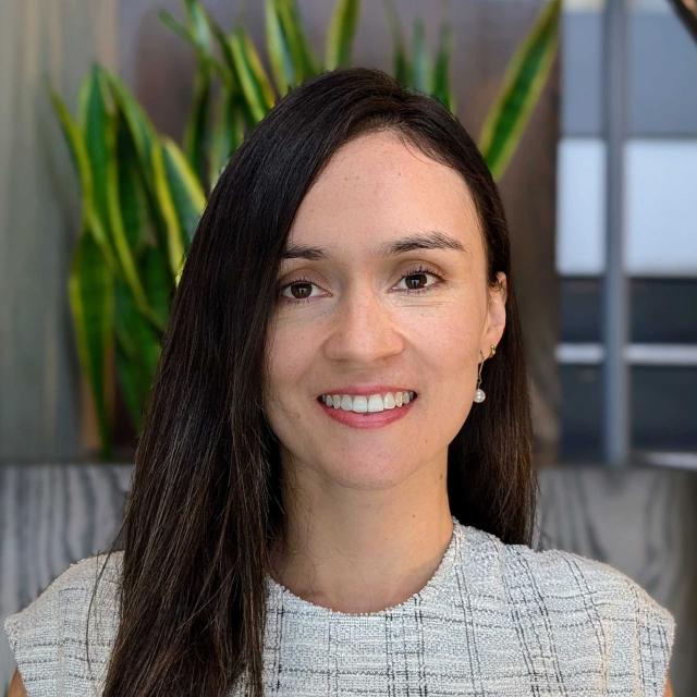 Professional headshot of a smiling woman wearing a white textured blazer, with indoor plants visible in the background