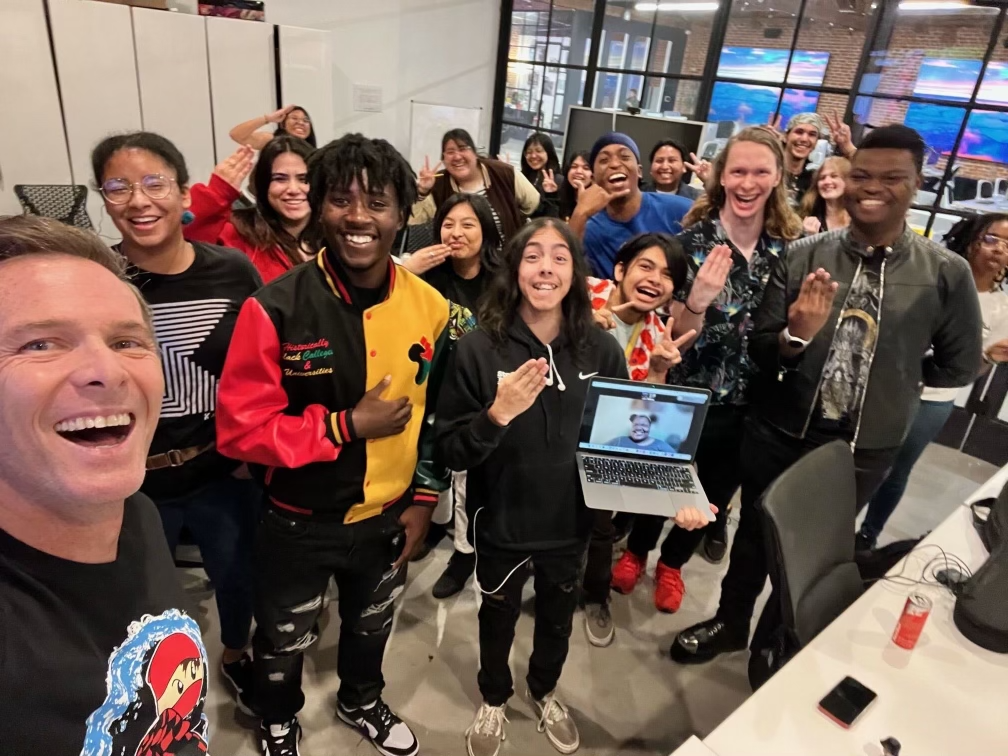 Large group of diverse students smiling and making celebratory gestures in a classroom, with one person joining virtually on a laptop