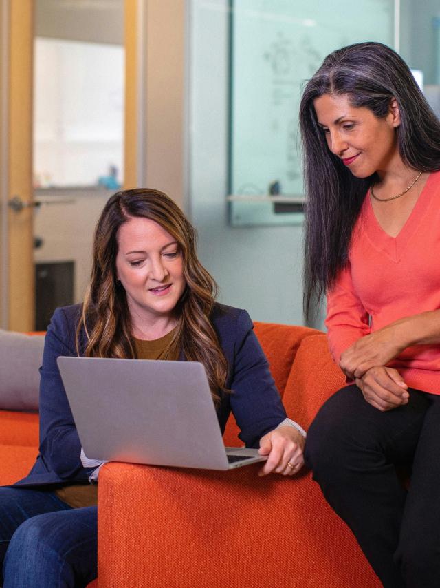 Two professional women having a casual meeting while reviewing work on a laptop, seated on a bright orange couch in a modern office setting