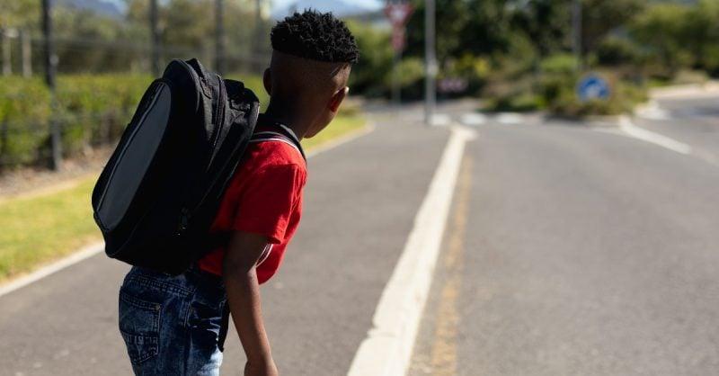 Side view of a student wearing a red t-shirt and black backpack walking along an empty rural road on a sunny day