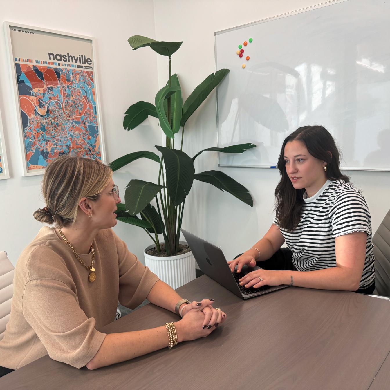 Two professionals having a meeting at a wooden table in a modern office space with a Nashville map poster and large indoor plant
