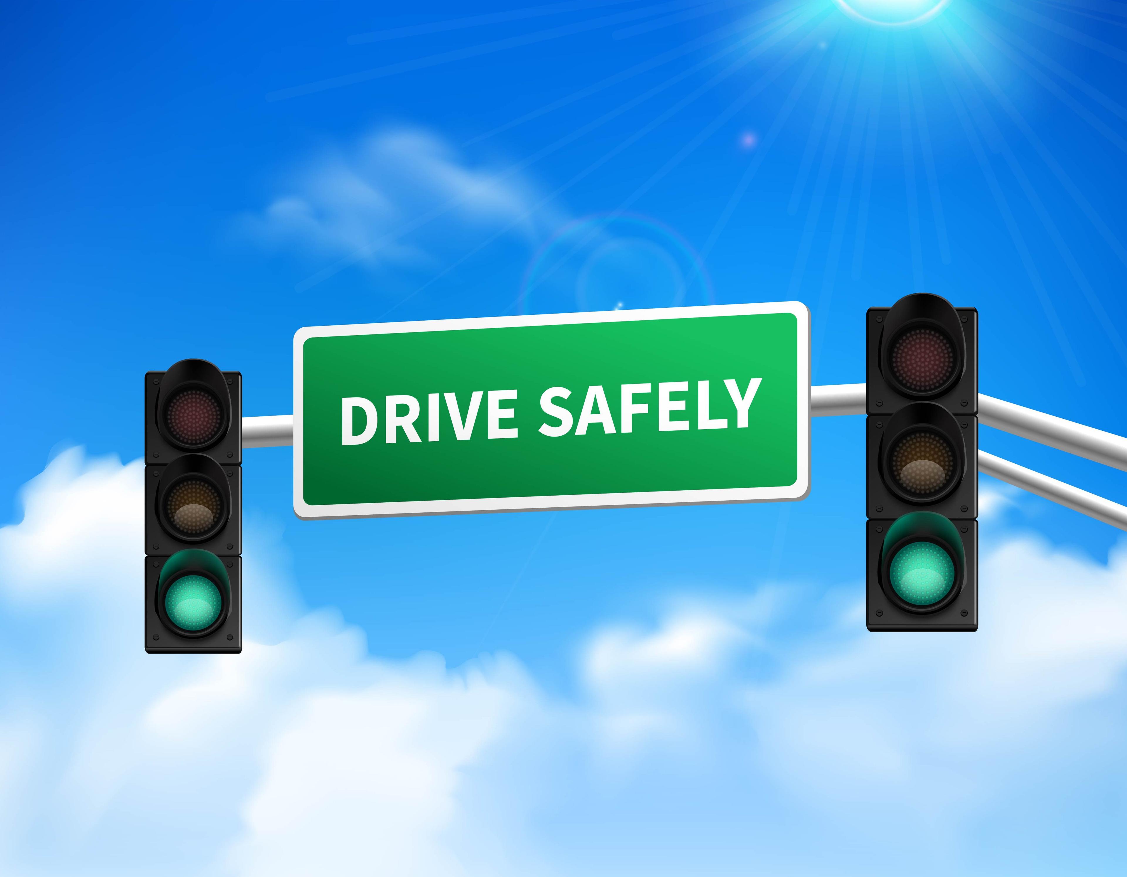 Green highway sign reading 'DRIVE SAFELY' mounted between two traffic lights showing green signals against a blue sky